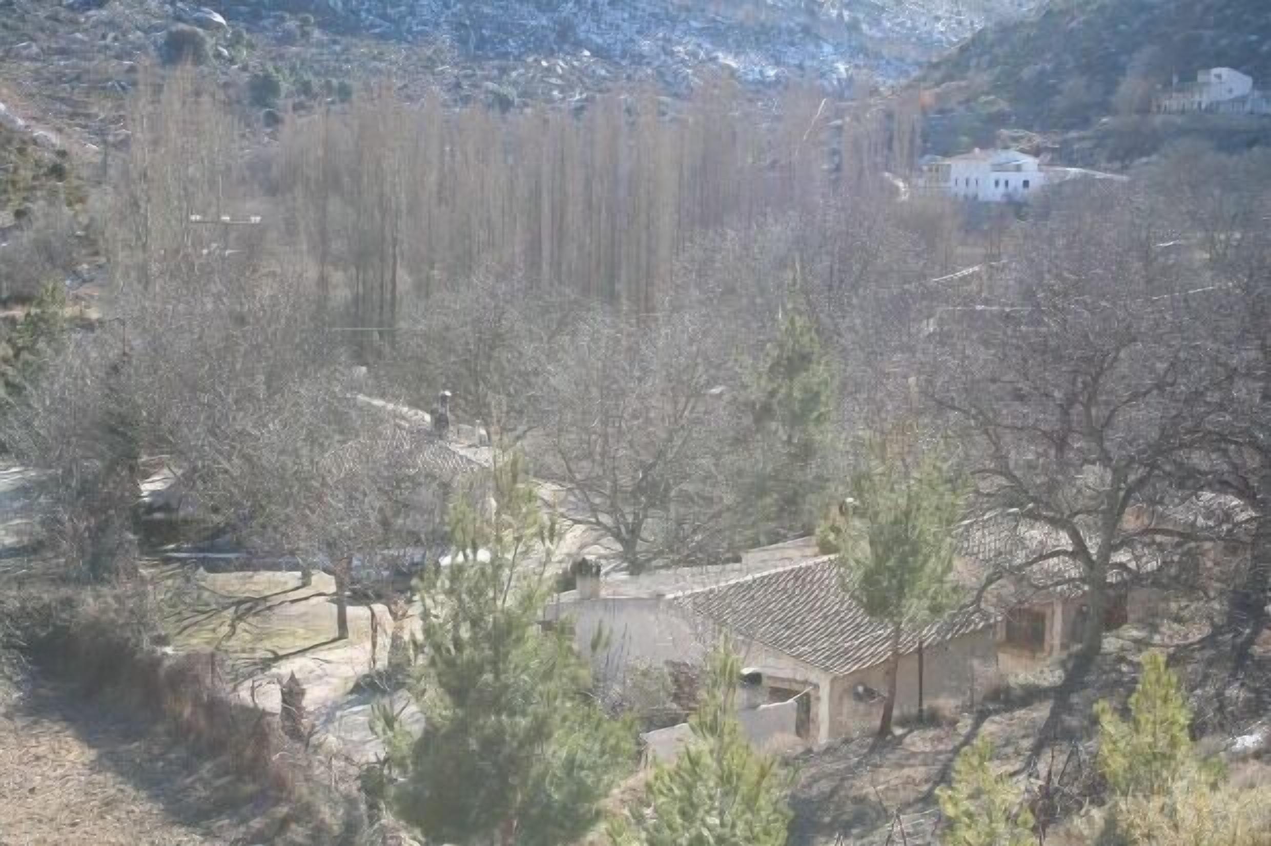 A mountainous landscape with sparse vegetation and a few buildings, including a white house with a tiled roof.