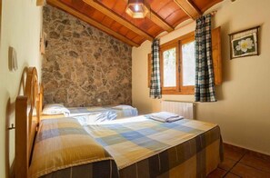 A bedroom with two beds, stone walls, wooden ceiling beams, a window with plaid curtains, and a flower painting on the wall.