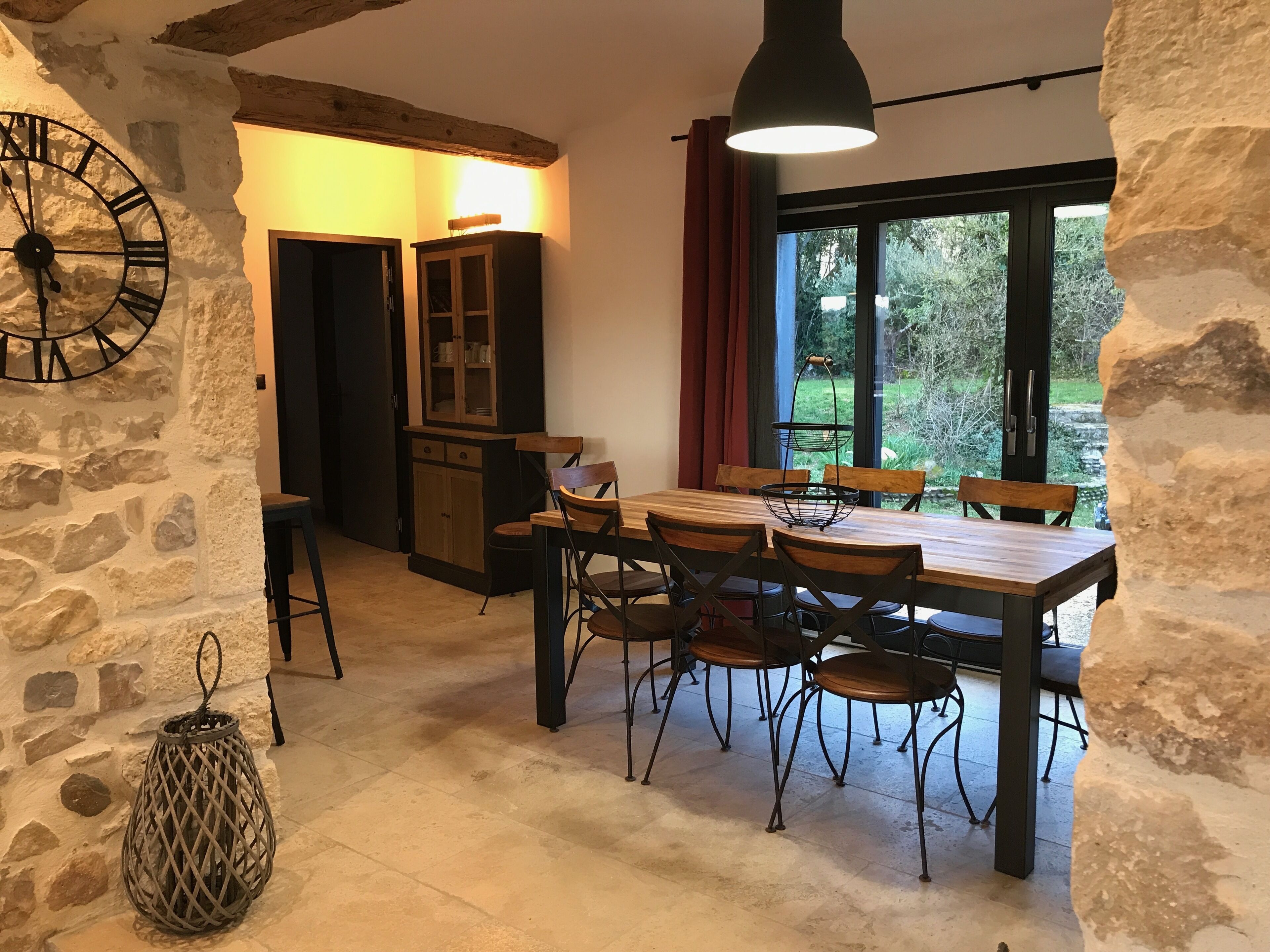A rustic dining area with a wooden table and chairs, a stone wall, a large clock, and a window with a view of greenery.