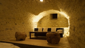 Ein gemütlicher, schummrig beleuchteter Raum mit einer Steinwand, einem Bett mit Kissen und einem kleinen Tisch.