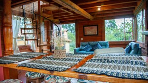 A wooden interior with a table covered by woven mats, a sofa with colorful cushions, and a window with a view of greenery.