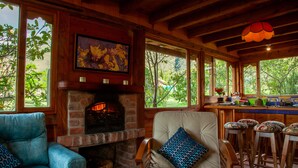 A cozy interior with a fireplace, wooden furniture, and large windows offering a view of greenery.