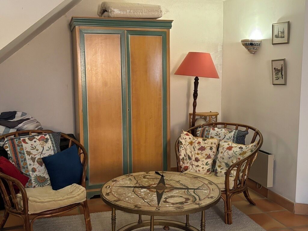 Un salon chaleureux avec un meuble en bois, des fauteuils en osier, une table ronde ornée d’une boussole, une lampe rouge et des tableaux encadrés accrochés au mur.