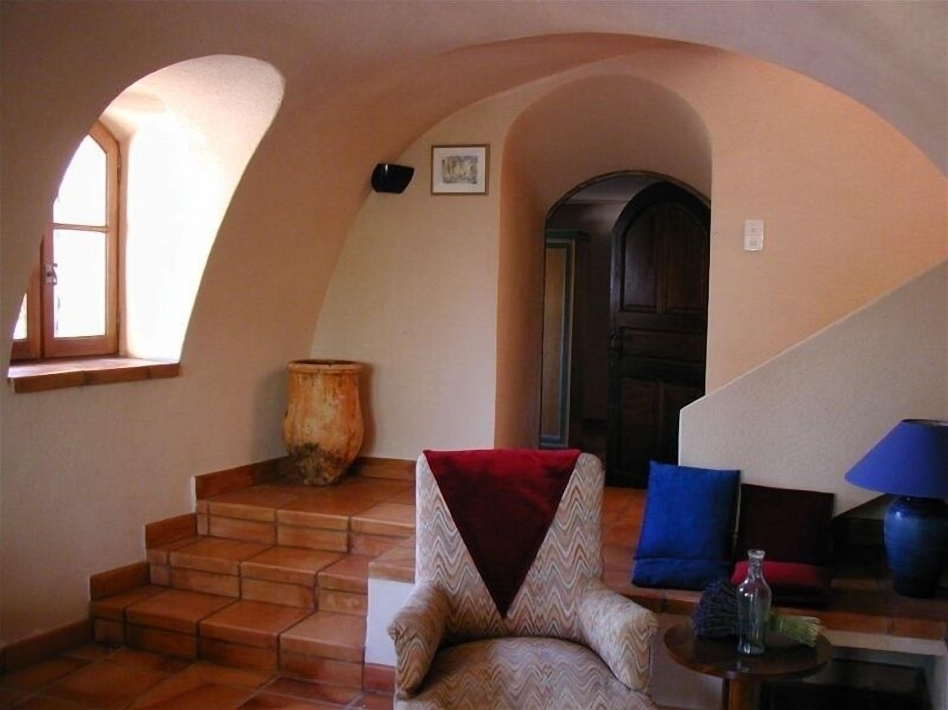 Un intérieur chaleureux avec un escalier menant à une porte sombre, une chaise dotée d’un coussin rouge et une lampe bleue posée sur une table.