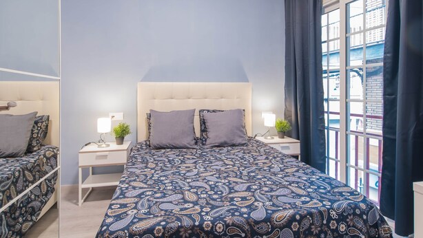 A bedroom with a large bed, two bedside tables with lamps, a nightstand with a plant, and a window with curtains. - DB Nice Apartment Sagrada Familia (Barcelona)