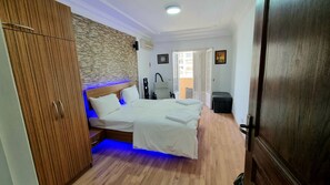 Un dormitorio con un armario de madera, una cama con ropa de cama blanca, una cabecera iluminada en azul y vistas al exterior a través de una puerta abierta.
