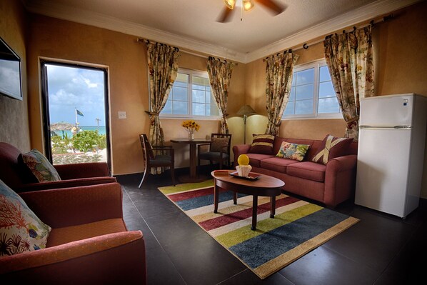 A living room with a sofa, armchair, coffee table, and a refrigerator. There is a window with curtains, a lamp, and a view of the outdoors.