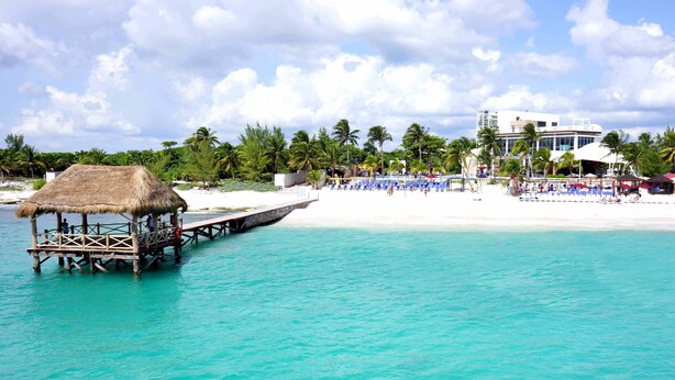 A beach with a thatched-roof structure extending into the water, a white sandy beach, and a building with a white facade in the background.