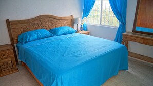 A neatly made bed with blue bedding, a wooden headboard with floral carvings, a bedside table with a lamp, and a window with blue curtains.
