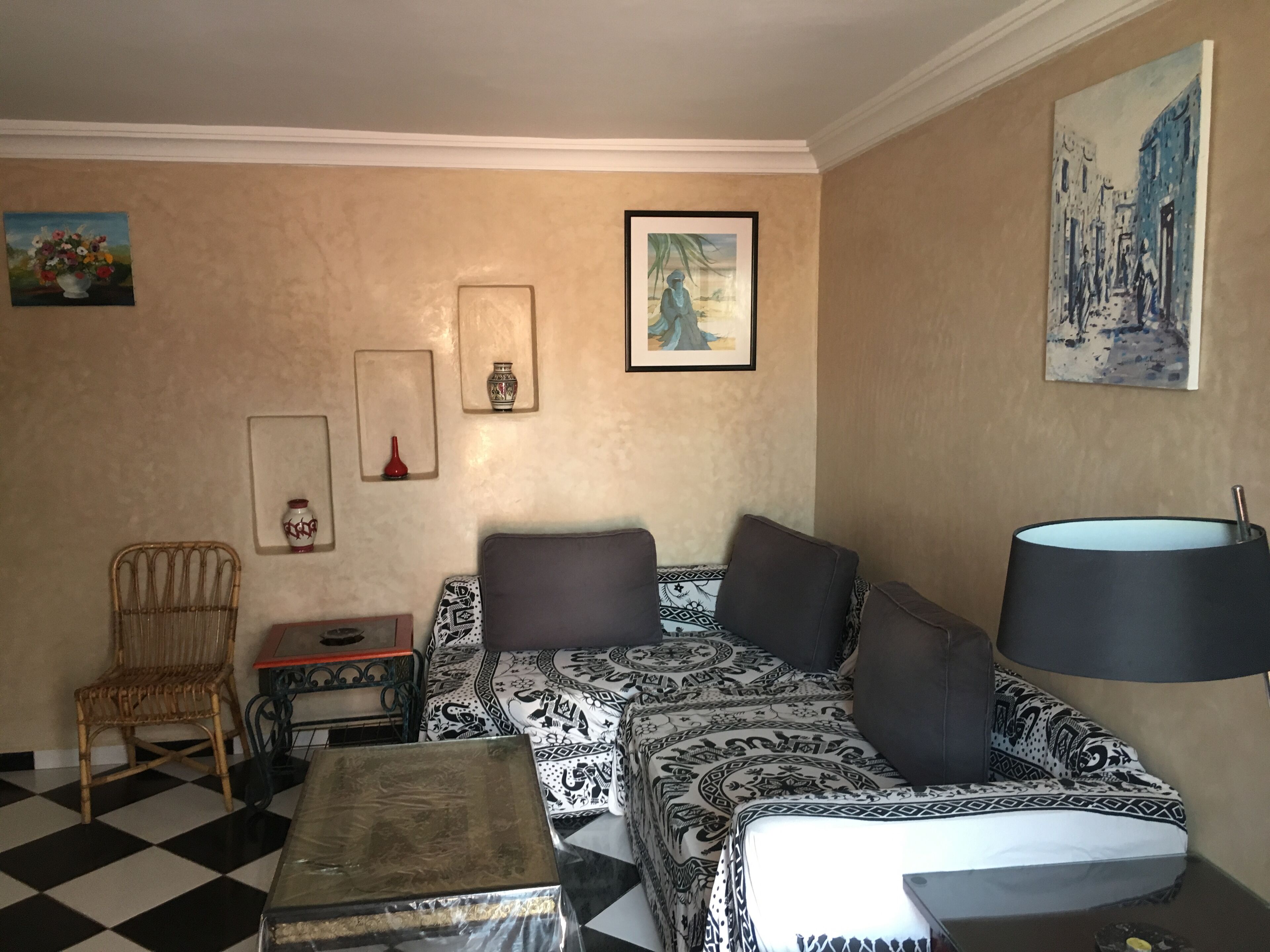 A cozy living room with a checkered floor, a sofa with patterned cushions, a small table, a chair, and wall art including a vase and a painting of a street scene.