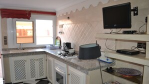 Una cocina con piso a cuadros blanco y negro, una ventana con cortina roja, estufa, lavaplatos y televisión montada en la pared.