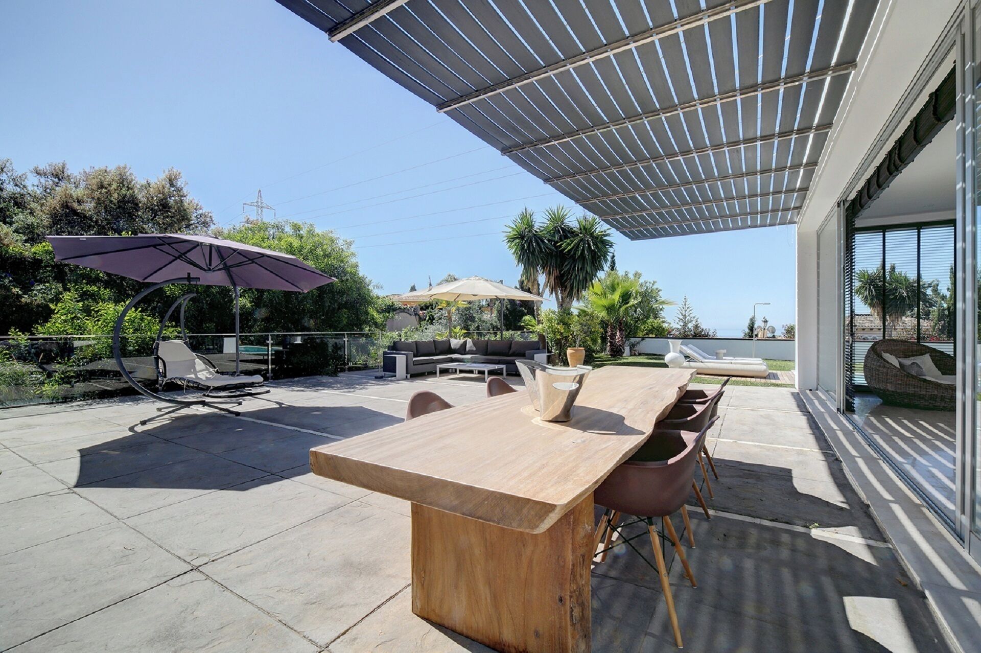 Een terras buiten met een houten tafel, stoelen en een pergola voor schaduw.