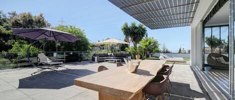 Een terras buiten met een houten tafel, stoelen en een pergola voor schaduw.