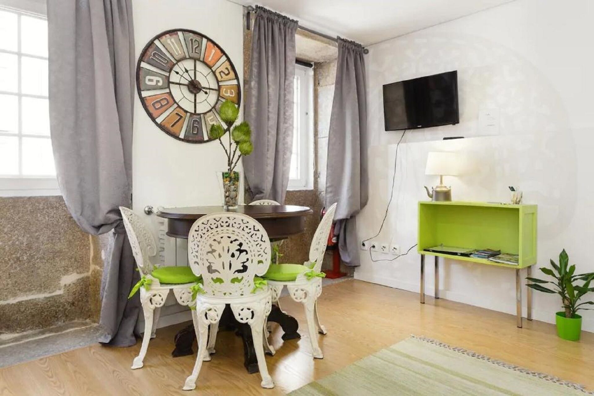 Un coin repas avec une table ronde, des chaises blanches, une horloge, un téléviseur et une étagère verte.