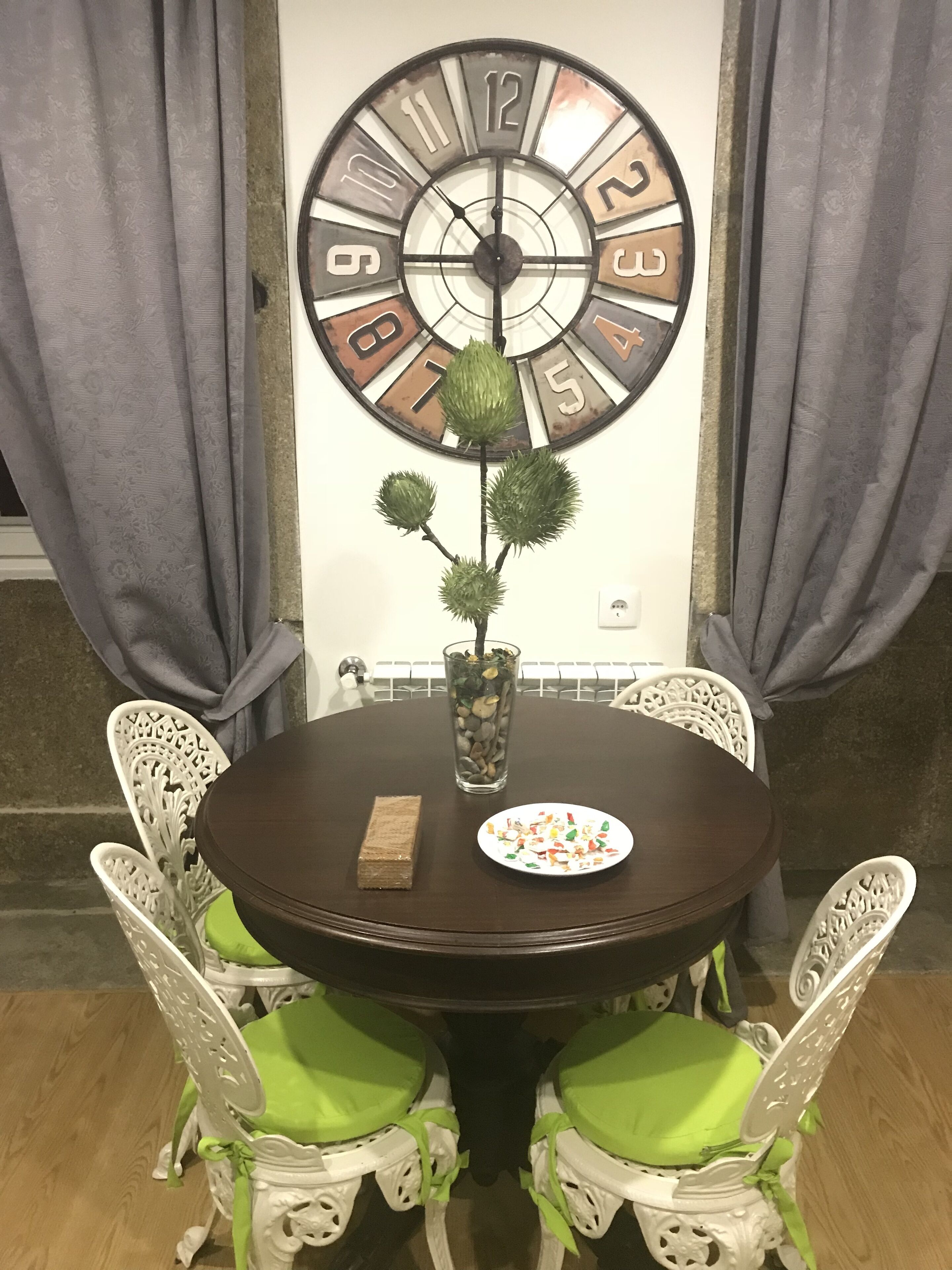 A dining table with a clock, chairs, a vase with a plant, and a plate of food.