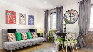 A living room with a grey sofa, colorful pillows, a round wall clock, and a dining area with a white table and chairs. - Home4you Downtown Porto (Porto)