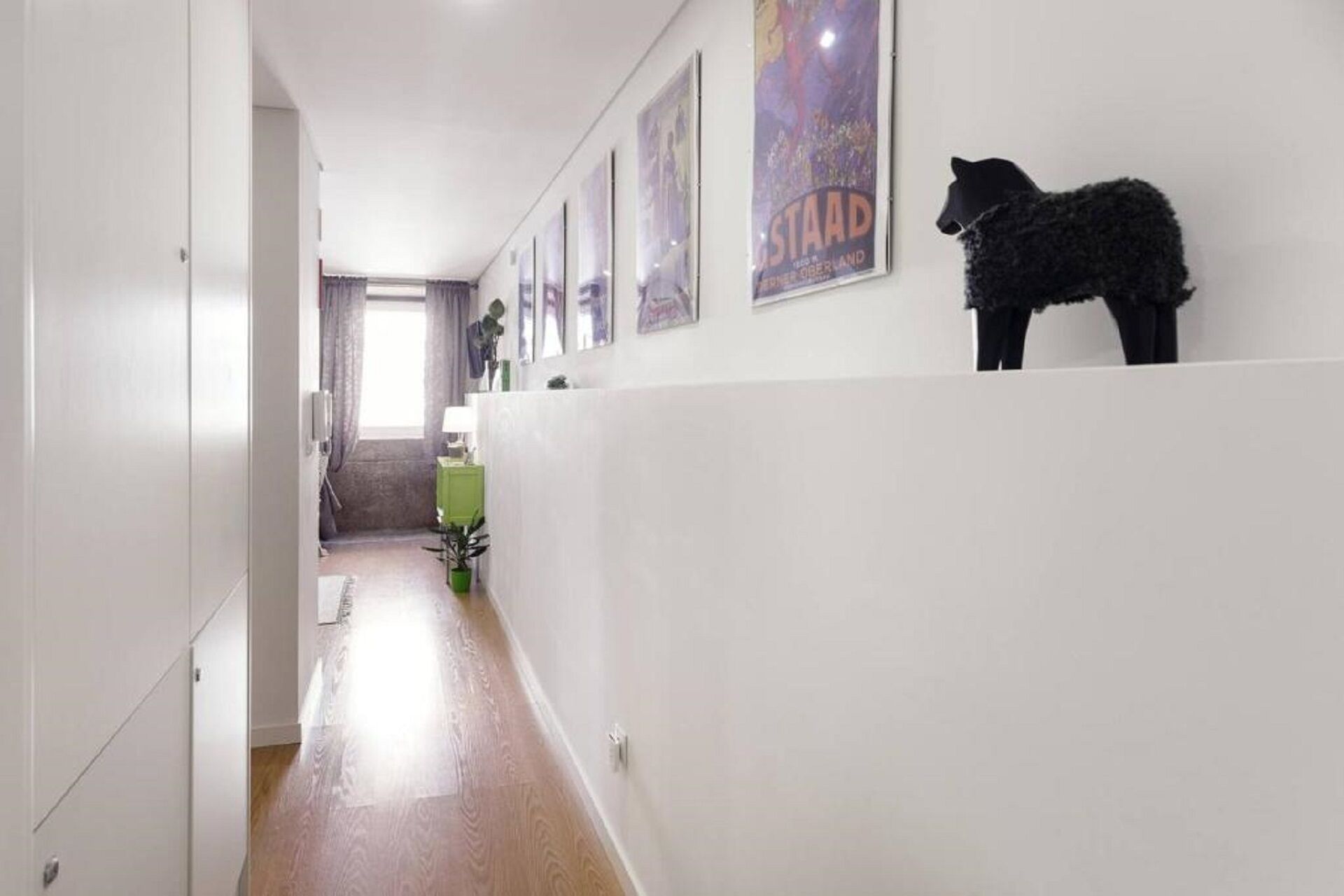 Un pasillo con piso de madera, paredes blancas y un armario verde. Hay carteles en la pared y un perro negro a la derecha.