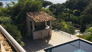 Ein Poolbereich mit Holzterrasse, einer kleinen Außenküche und Blick auf Meer und Berge.
