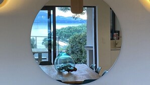 Ein Essbereich mit Tisch und Stühlen, ein großes Fenster mit Blick auf ein Gewässer und Berge sowie ein runder Spiegel an der Wand.
