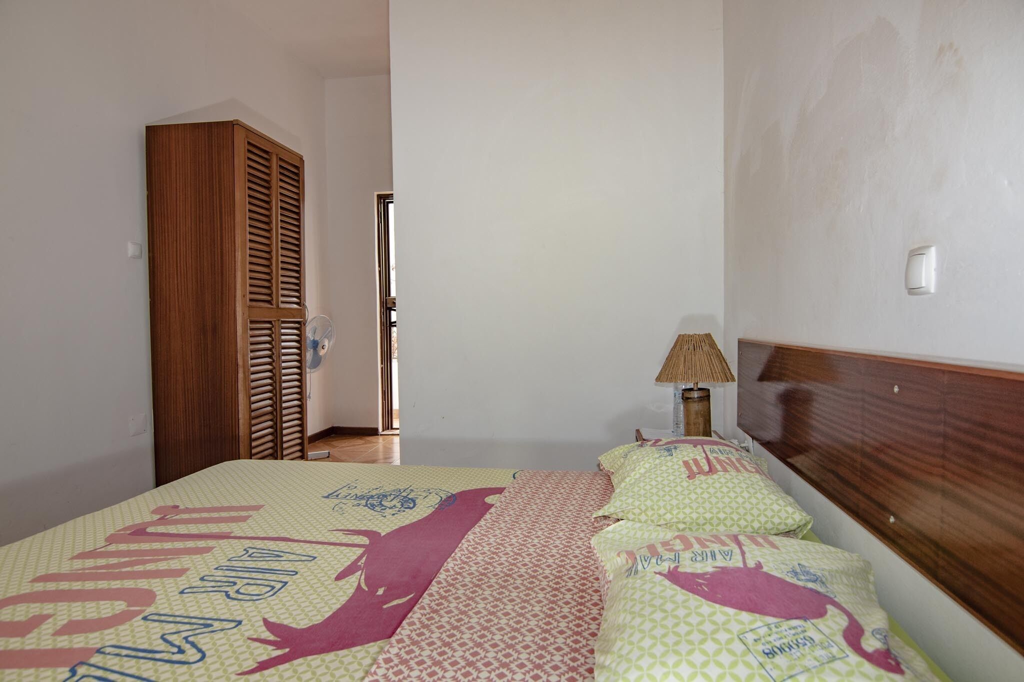 Une chambre à coucher avec un lit, une armoire en bois, un ventilateur sur pied et une lampe de chevet.