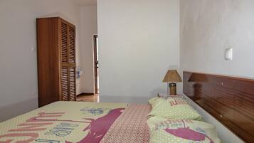 Une chambre à coucher avec un lit, une armoire en bois, un ventilateur sur pied et une lampe de chevet.
