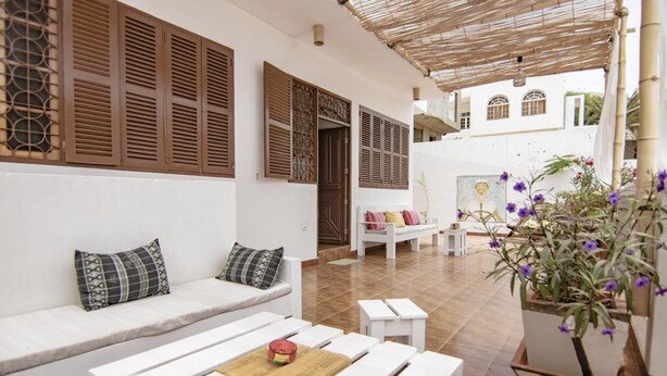 A white outdoor seating area with wooden benches and cushions, a woven ceiling, and a potted plant with purple flowers. - Holiday house 200 meters from Laginha beach (Mindelo)