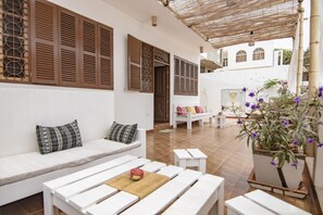 A white outdoor seating area with wooden benches and cushions, a woven ceiling, and a potted plant with purple flowers. - Holiday house 200 meters from Laginha beach (Mindelo)