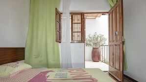A bedroom with a bed, a window with curtains, a balcony with a potted plant, and a wooden door. - Holiday house 200 meters from Laginha beach (Mindelo)