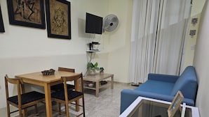 Uma sala de estar com uma mesa de jantar, cadeiras, um sofá azul, uma mesa de centro de vidro, uma televisão, um ventilador e uma planta.
