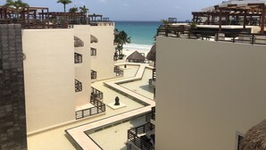 Un resort con vista al océano, que cuenta con un área de piscina con diseño geométrico, asientos en terraza de madera y una estructura con techo de paja.