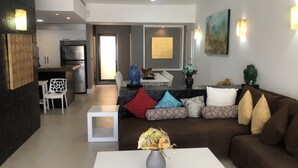 Una sala de estar moderna con un sofá marrón, una mesa de centro blanca y un jarrón con flores secas. Hay un comedor con una mesa y sillas blancas, y una cocina con un refrigerador y armarios.