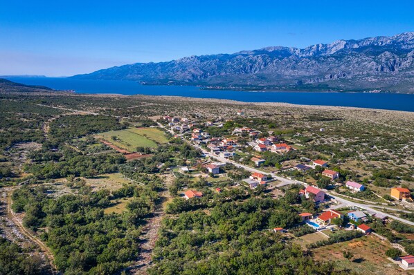 Vista aérea de un pueblo costero con casas, una carretera y una extensa masa de agua con montañas al fondo.