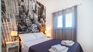 Hotelkamer met een groot bed, twee nachtkastjes, een muurschildering van een stadsgezicht, een raam met gordijnen en een lamp op elk nachtkastje.