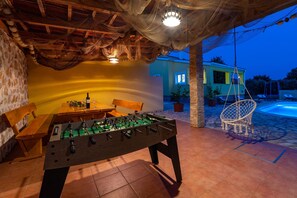 Una mesa de fútbol de mesa en un patio exterior con un banco y mesa de madera, una silla colgante y una piscina al fondo.