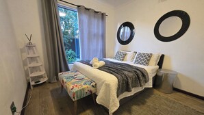 Ein Schlafzimmer mit Bett, einer kleinen Bank, einem Regal, einem Fenster mit Vorhängen und einem an der Wand befestigten Spiegel.