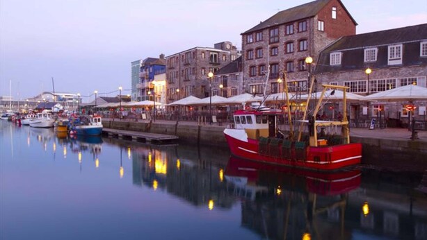 A harbor with boats docked, buildings with multiple stories, and outdoor seating areas with umbrellas. - Quirky Coach House on Plymouth Hoe (Plymouth)