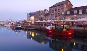 A harbor with boats docked, buildings with multiple stories, and outdoor seating areas with umbrellas. - Quirky Coach House on Plymouth Hoe (Plymouth)