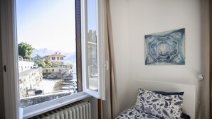 A bedroom with a bed, a radiator, a window with a view of a building and mountains, and a painting on the wall.