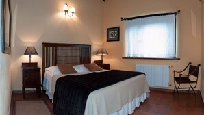 Un dormitorio con una cama, dos mesitas de noche con lámparas, una silla, un ventanal con cortinas y un radiador.