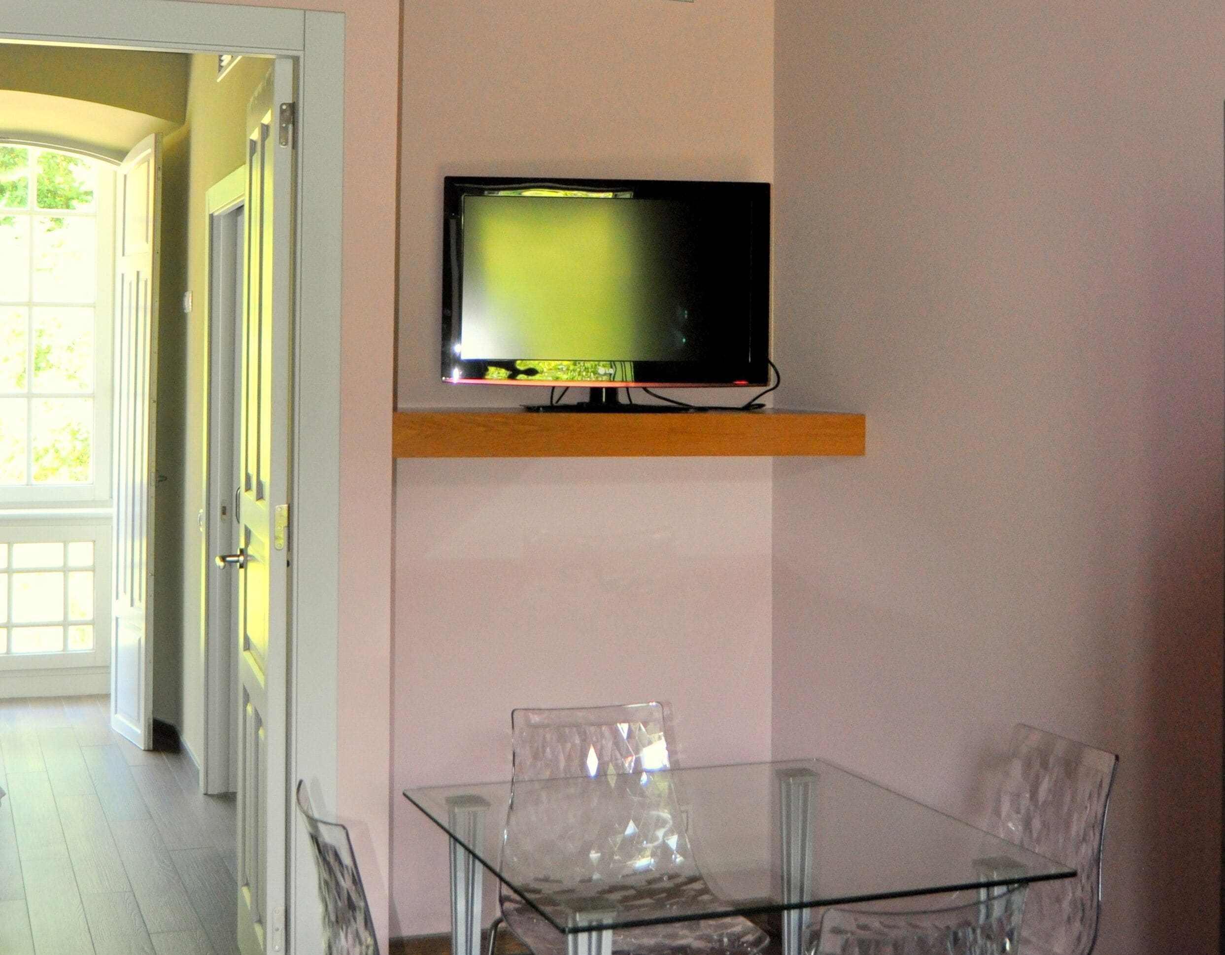 Una habitación con una mesa y sillas de vidrio, un televisor montado en la pared y una puerta que da a un pasillo luminoso.