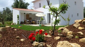 A white house with a tiled roof, a patio with outdoor furniture, and a garden with red flowers and rocks. - Casa do Olival, Guest accommodation in Figueiró dos Vinhos. (Figueiró dos Vinhos)