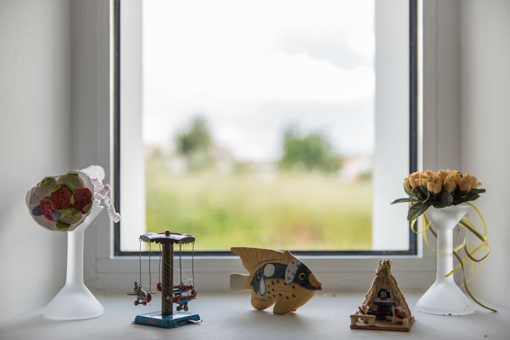 A window with a view of a green field, a white vase with a bouquet of flowers, a small wooden boat, and a toy carousel.