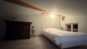 A bedroom with a wooden beam ceiling, a bed, a small table with a lamp, and a window.