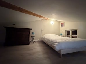 A bedroom with a wooden beam ceiling, a bed, a small table with a lamp, and a window.