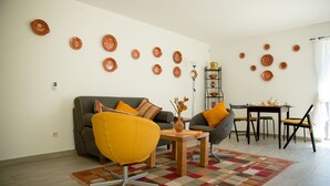 Una sala de estar con un sofá, dos sillones, una mesa de centro y una mesa de comedor con sillas. Hay platos decorativos en la pared y un jarrón con flores secas sobre la mesa de centro.