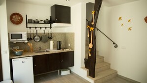 Cocina compacta con armarios oscuros, microondas y lavamanos. Hay un refrigerador pequeño y una encimera con diversos utensilios. Una escalera de madera con un diseño único conduce a otro nivel.