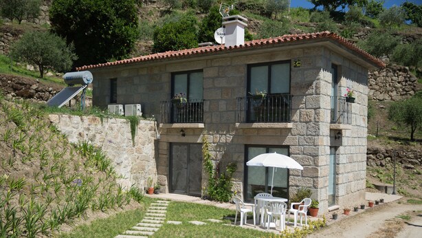 Una casa de piedra con techo de tejas, un balcón con barandilla de metal, un pequeño jardín con plantas en macetas y un paraguas blanco.