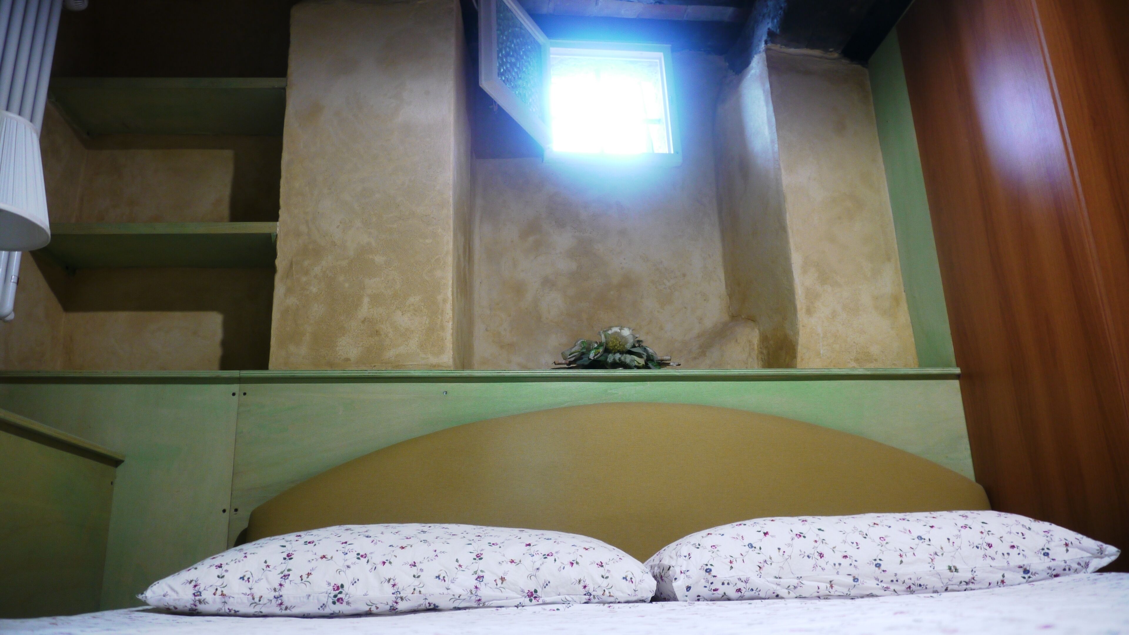 Un dormitorio con una cama, almohadas, una mesita de noche, un ventanal y un estante en la pared.