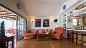 Un salon comprenant un canapé, des fauteuils, une table basse et un téléviseur posé sur une table en bois. Il y a également un coin cuisine avec des tabourets de bar et une fenêtre.