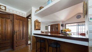 Une cuisine avec une porte en bois, un plan de travail blanc, deux tabourets de bar, un saladier rempli de fruits et un réfrigérateur.